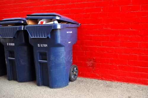 Two large blue plastic wheelie bins with black lids are positioned side by side on a paved outdoor surface, directly in front of a red brick wall. The bins are slightly tilted forward, revealing some visible waste materials inside the partially open lids. The surface appears to be concrete or asphalt, with small gravel particles scattered around. The bins have textured surface patterns and are equipped with wheels on their rear side for mobility. The red brick wall behind them is painted uniformly, providing a contrasting background that emphasizes the bins' presence. This scene is typical of commercial waste disposal outside a building, reflecting rubbish collection practices often handled by companies like Commercial Waste Havering, serving the Havering area in the UK, where efficient rubbish management is essential for local businesses and properties.
