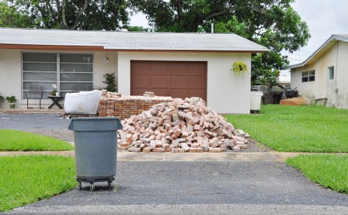 A pile of light-colored bricks and rubble, consisting of broken brick fragments and small stone pieces, is situated on the driveway in front of a single-story residential building with a white exterior wall, large window, and brown garage door. To the left of the pile, a grey wheeled rubbish bin with a dark lid is positioned on the asphalt surface. The scene is set on a well-maintained suburban street with bright green grass patches bordering the driveway and surrounding the house. In the background, the house features a window with horizontal blinds and a small potted plant hanging on the wall near the garage. The sky appears overcast, providing soft, diffused lighting. This image reflects the sort of scene where a professional rubbish removal service, such as Commercial Waste Havering, might be involved in clearing waste materials from a residential property in Havering or nearby east London areas.
