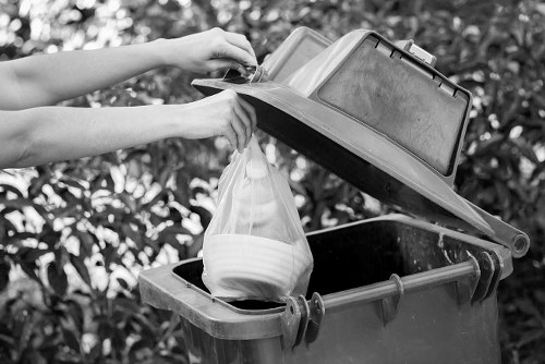 A person is seen disposing of waste into a black outdoor rubbish bin, which has a partially open lid revealing a plastic bag filled with mixed household waste. The individual is holding a small rectangular container or lid, poised above the bin's opening, with their arms extending from the left side of the image. The bin is situated on a paved surface adjacent to greenery with dense foliage, suggesting an outdoor setting in a residential or commercial area within Havering. The bin appears to be made of durable plastic, with a textured surface in a dark color, supported by a metal handle. The scene is captured in natural daylight, providing clear visibility of the bin's contents and surroundings. This image relates to rubbish disposal or waste management services, such as those offered by Commercial Waste Havering, and visually emphasizes proper waste disposal practices in the local area.