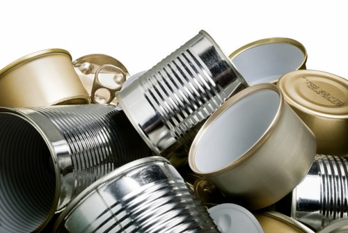 A collection of discarded metal cans, some with pull-tab openings and others with plain lids, piled together in a mixed heap. The cans exhibit a variety of finishes, including shiny silver and matte gold, with some showing embossed branding or labels. They are positioned in a disorganized manner, with some lying on their sides and others stacked or leaning against each other, on a flat surface that appears to be outdoors or in a storage area. The background is blurred, emphasizing the focus on the cans' metallic textures and cylindrical shapes. The scene reflects the type of waste material that services like Commercial Waste Havering handle for rubbish removal, particularly metal packaging waste common in refurbishment or commercial disposal contexts around Havering, London postal code area. The lighting is natural, highlighting the reflective surfaces and metallic hues, contributing to a clean and detailed visual description suited for accessibility and local SEO relevance.