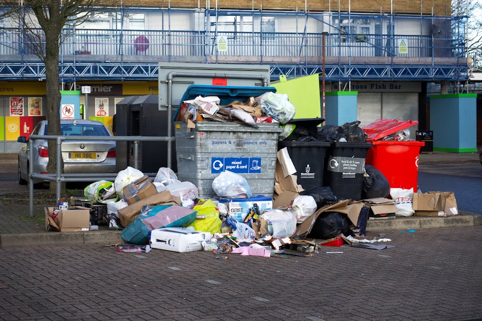 A waste collection worker wearing a red and yellow uniform is seen operating a large red rubbish truck on a street, with the truck's rear hatch open for loading debris. The worker is standing on the g