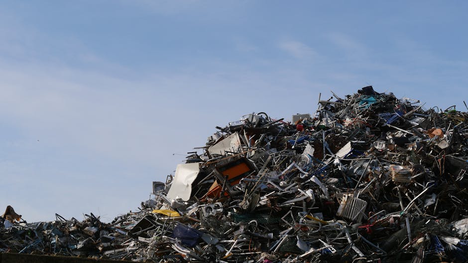 A large, untidy pile of mixed metal, plastic, and wooden debris sits outdoors against a backdrop of clear blue sky. The rubbish includes metallic appliances, broken furniture, scattered wires, and mis