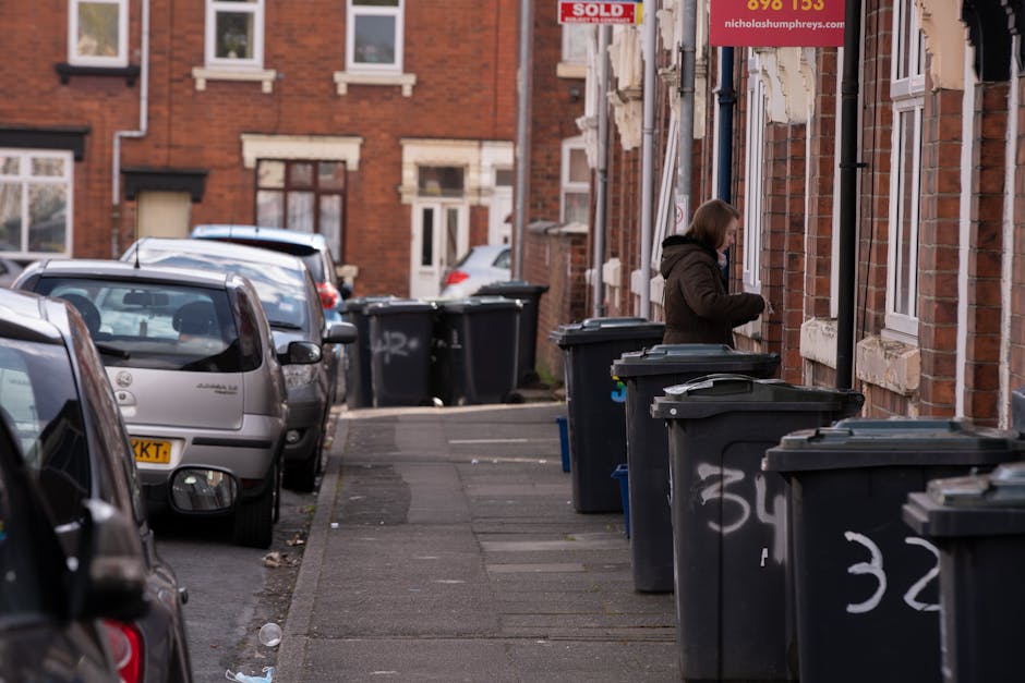 A sanitation worker wearing a blue uniform and an orange reflective vest is placing a blue wheelie bin into the rear compartment of a white waste collection truck parked on a narrow urban street. The 